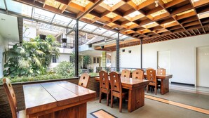 A spacious indoor area with a long wooden table and chairs, large windows, and a view of a garden.