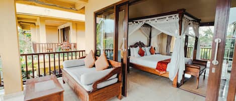 A room with a bed, a sofa, and a small table. There is a balcony with a railing and a view of the outside.