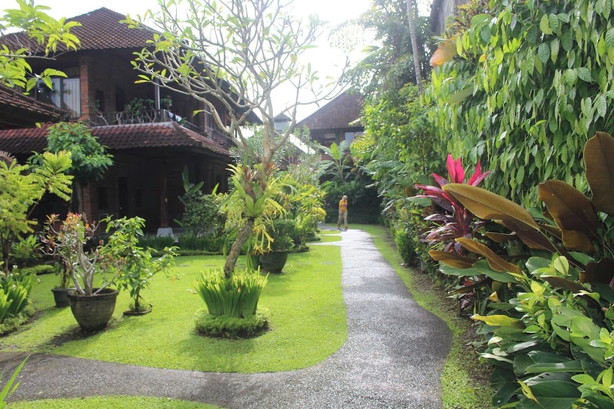 A pathway leading through a lush garden with various plants and a traditional building with a tiled roof.
