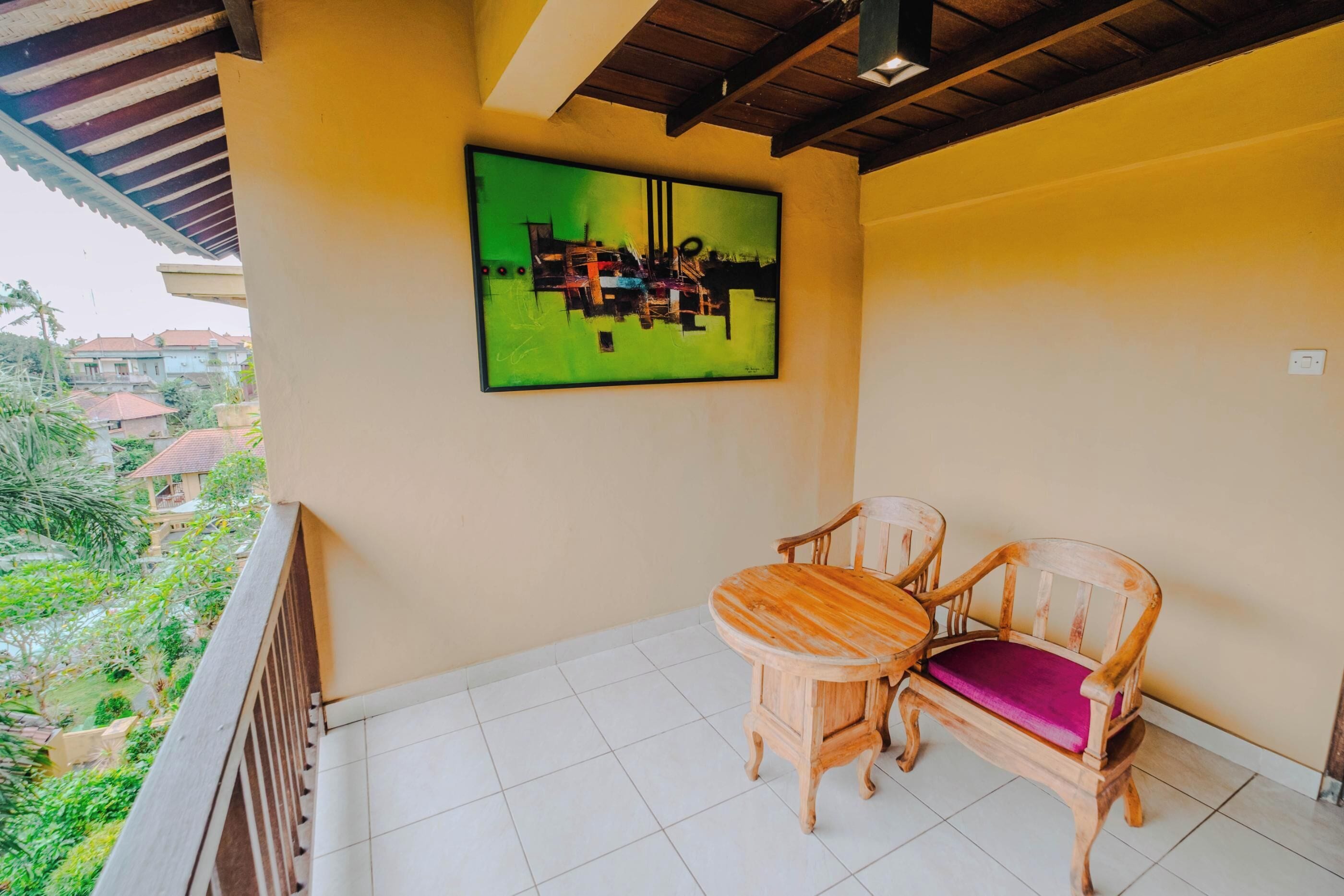 A balcony with a wooden table and chairs, a green abstract painting on the wall, and a view of greenery and buildings in the background.
