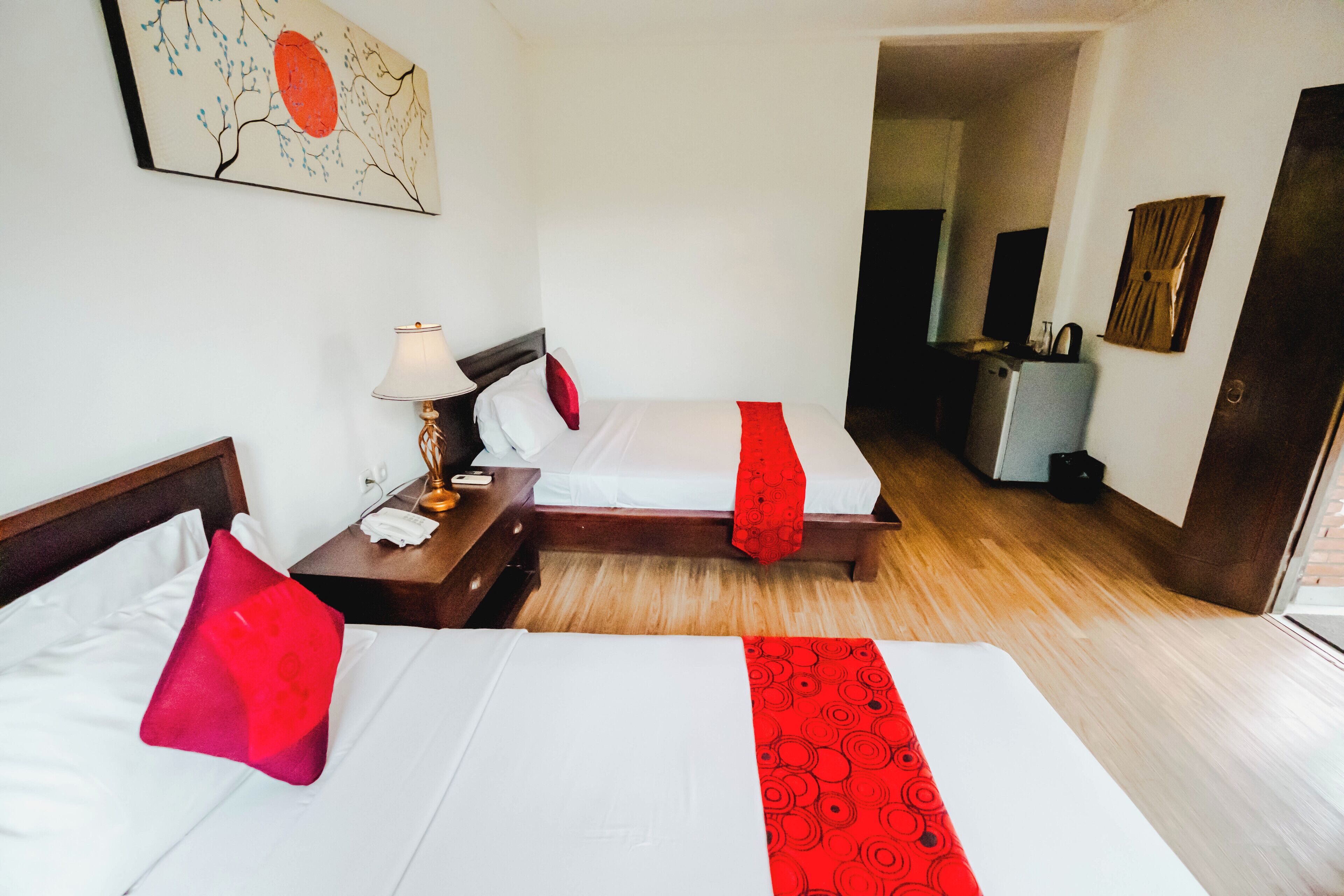 Habitación de hotel con dos camas, una mesita de noche, una lámpara, un cuadro y una puerta que da a otra habitación.