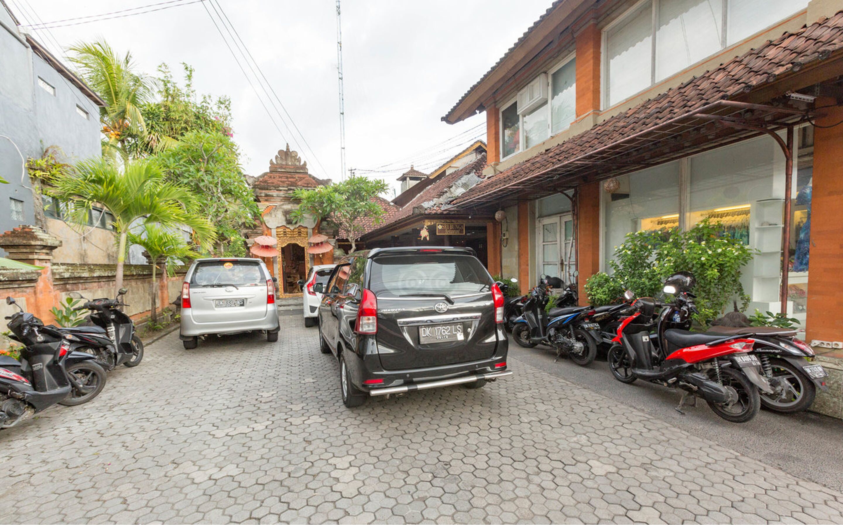 Uma rua com carros e motocicletas estacionados, um edifício tradicional com telhado de azulejos e uma placa com a inscrição "Sukomal".
