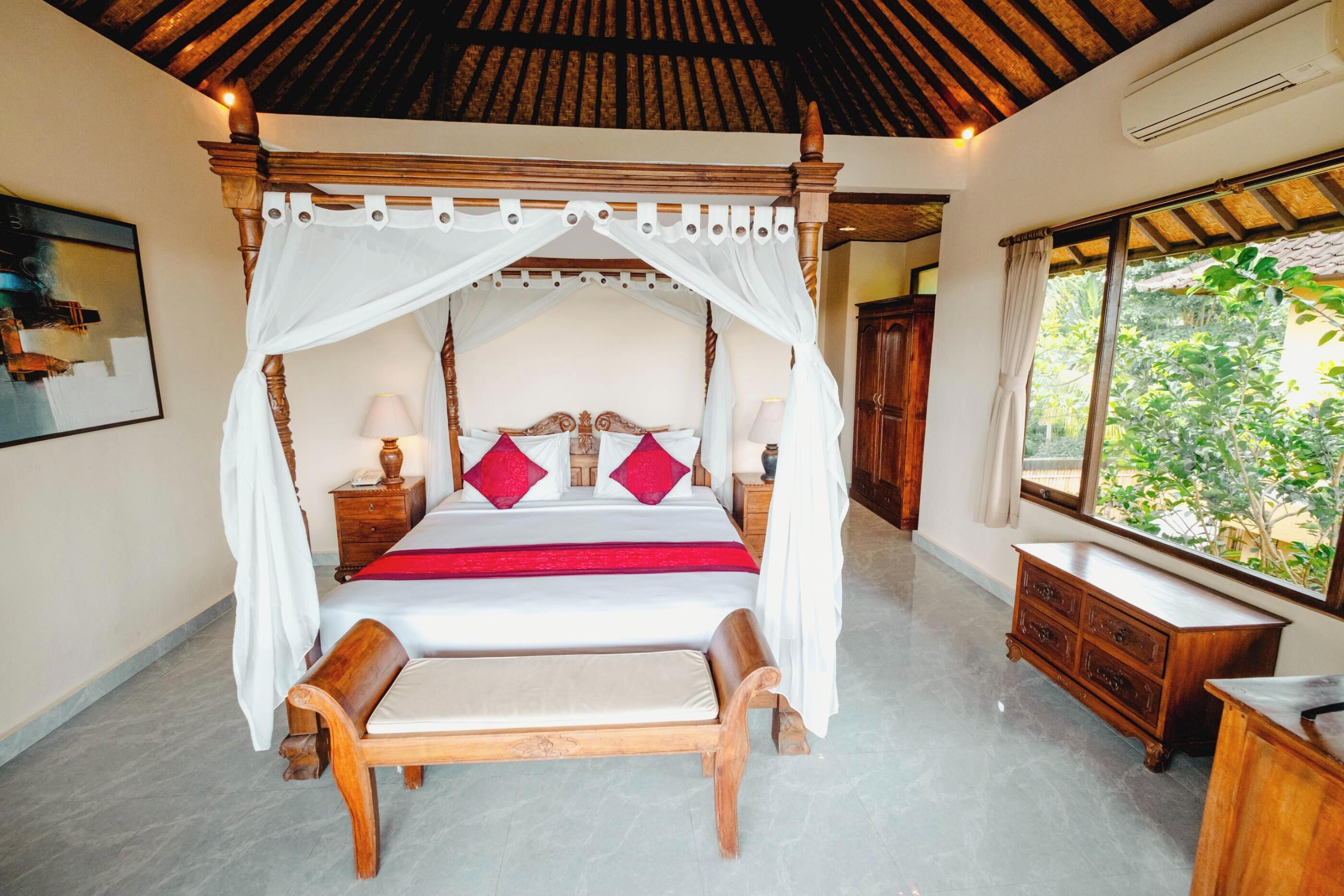 A four-poster canopy bed with a wooden frame, a red bedspread, and two red pillows. There is a wooden nightstand with a lamp, a wooden dresser, and a window with a view of greenery outside.