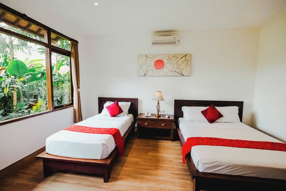 A hotel room with two beds, a nightstand with a lamp, and a window with a view of greenery.