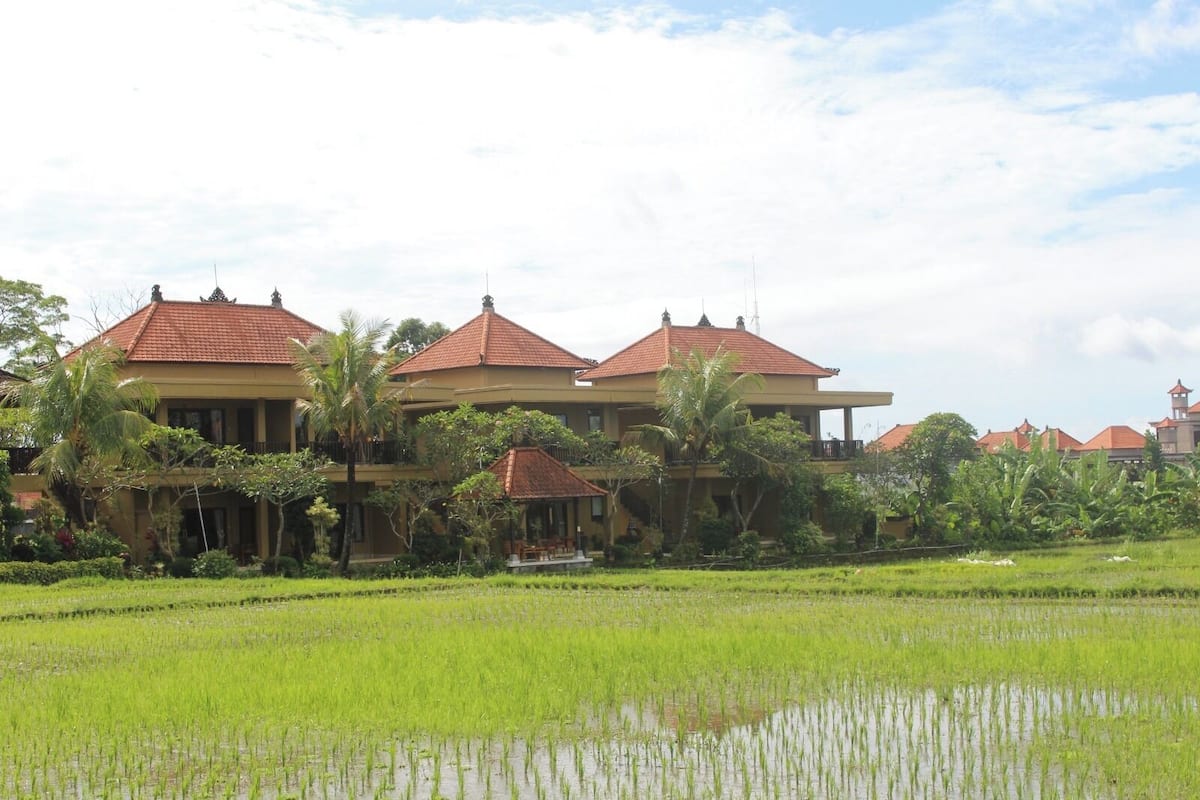 A two-story building with a red-tiled roof, surrounded by lush greenery and a rice field.