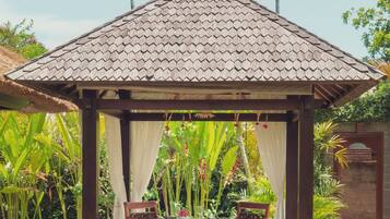 Um gazebo com telhado ladrilhado, cortinas brancas e uma mesa com cadeiras.