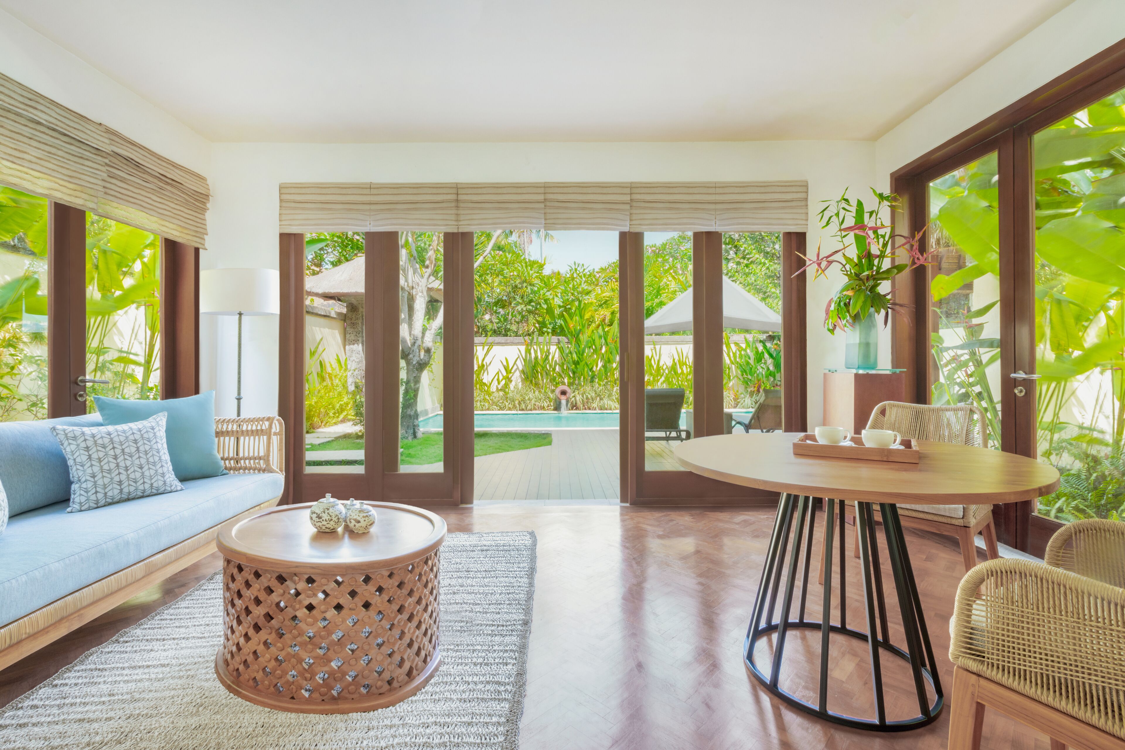 Una sala de estar moderna con sofá, mesa de centro y sillas, ventanas amplias con persianas y vistas a un jardín y piscina.