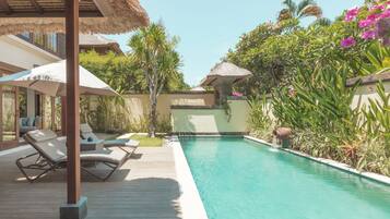 A swimming pool with lounge chairs and umbrellas, surrounded by tropical plants and a thatched roof structure.