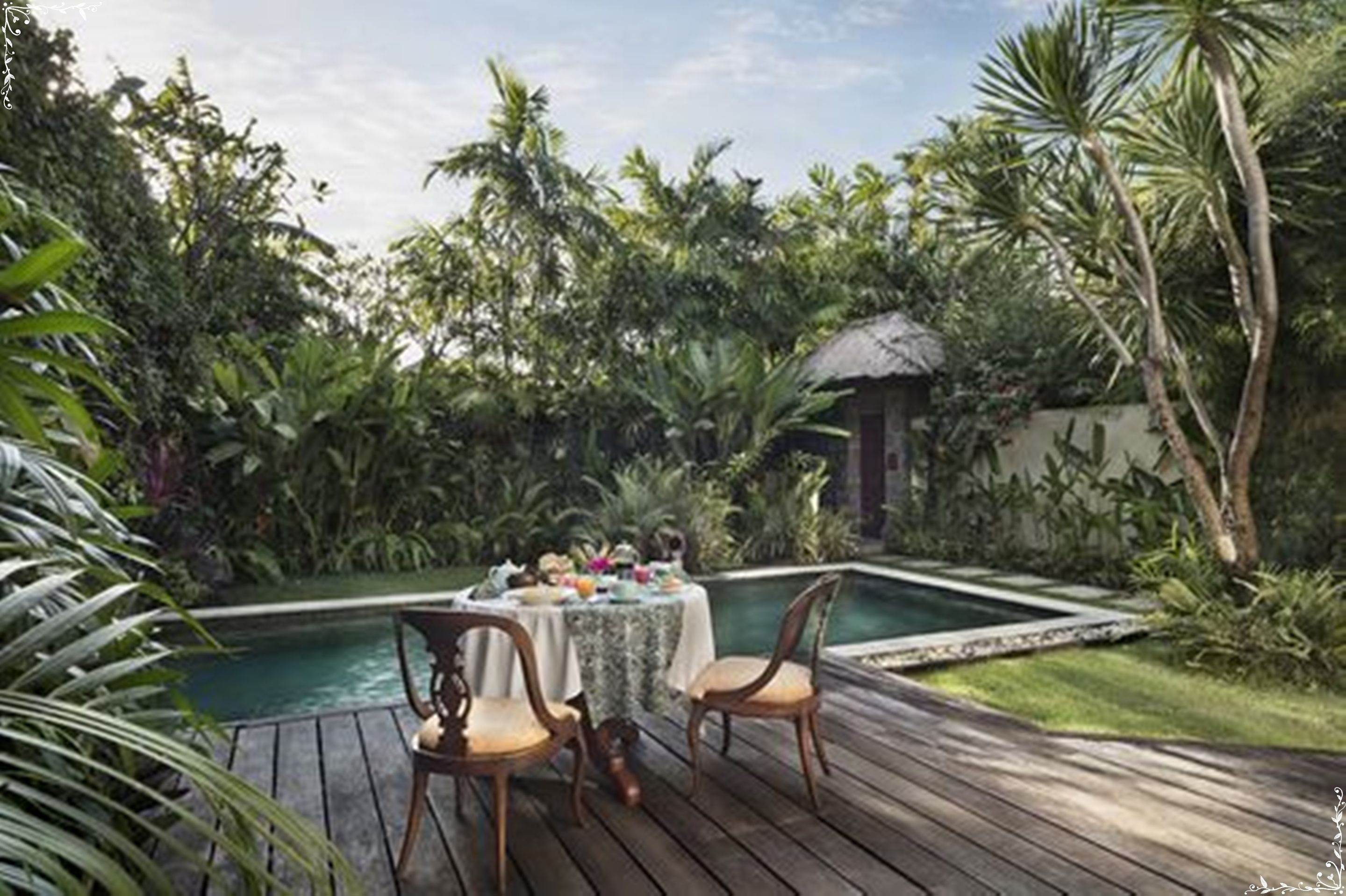 Un área de comedor junto a la piscina con una mesa para dos, rodeada de exuberante vegetación tropical.