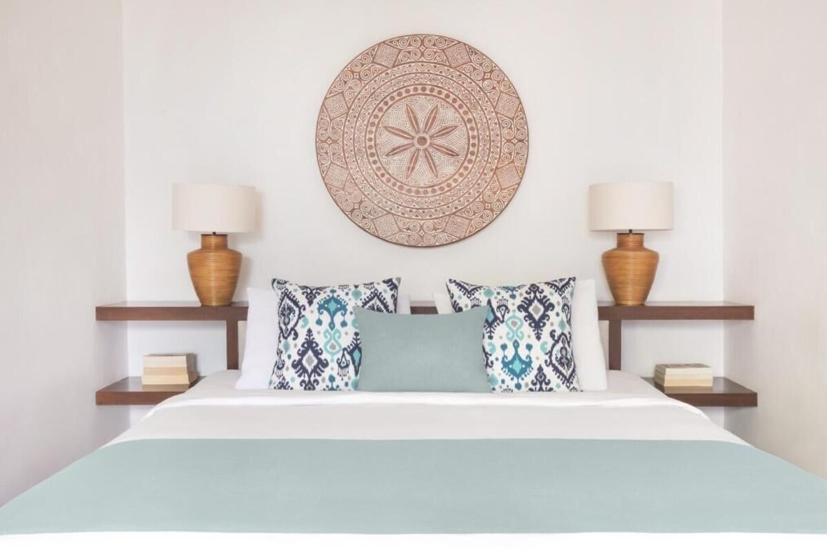 A bed with patterned pillows, a round wall decoration, and two bedside lamps on wooden nightstands.