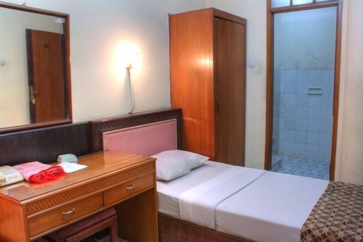 A hotel room with a wooden desk, a bed with white bedding, a wardrobe, and a bathroom with a tiled shower.