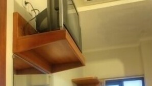 Une pièce de taille modeste, avec un bureau en bois, une chaise et un téléviseur posé sur une étagère. Il y a des fenêtres et une porte.
