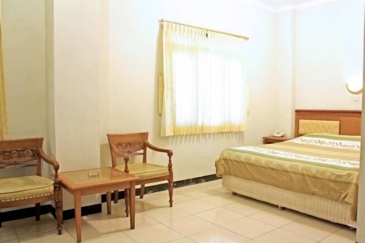 A hotel room with a bed, a wooden chair, a small table, and a window with curtains.