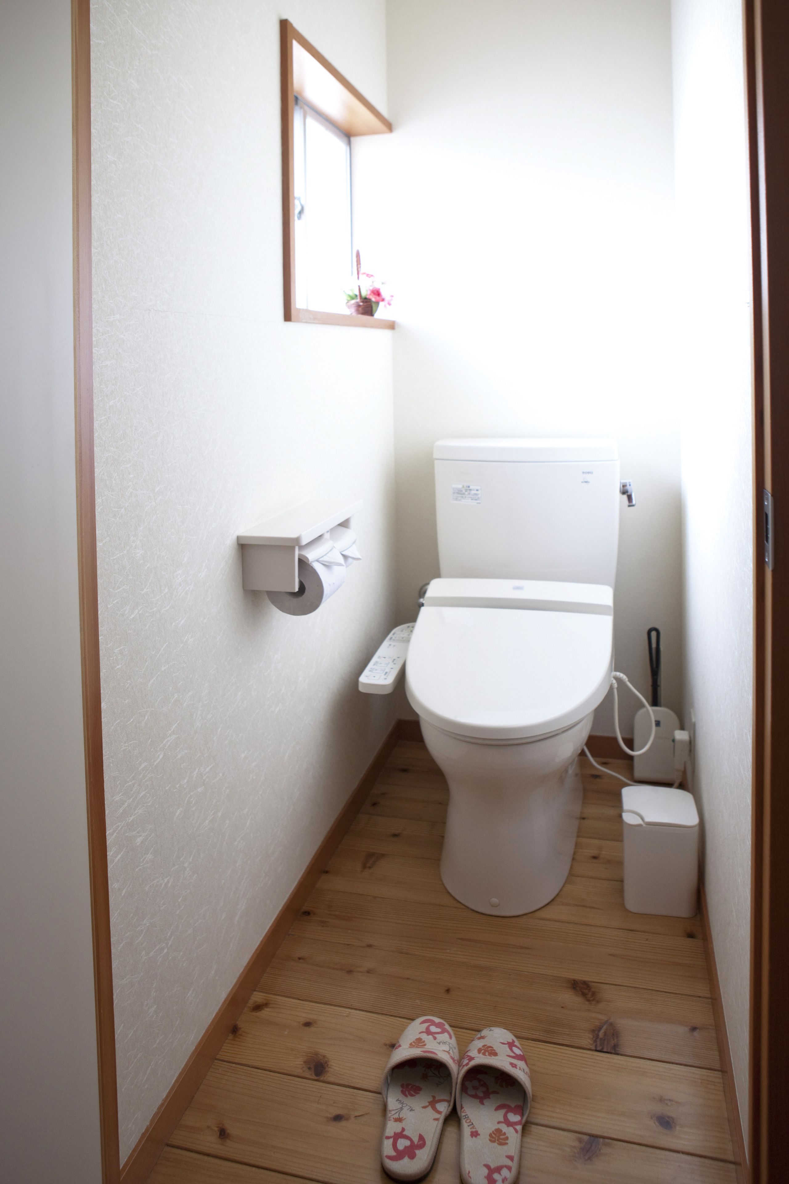 A small, narrow bathroom with a toilet, a small shelf with a tissue box, and a pair of slippers on the floor.