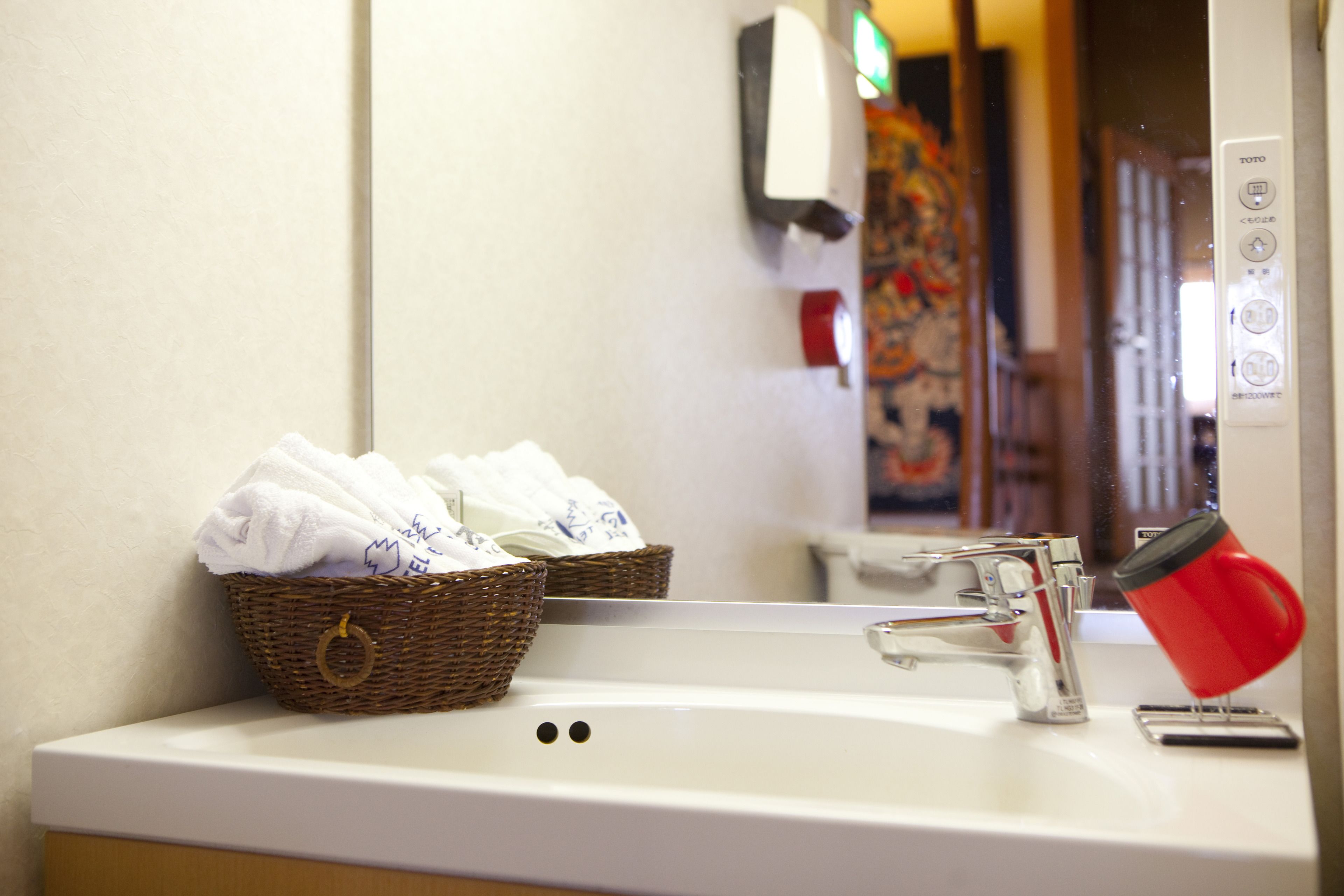 A bathroom sink with a basket of towels, a red cup, and a mirror reflecting a colorful painting.