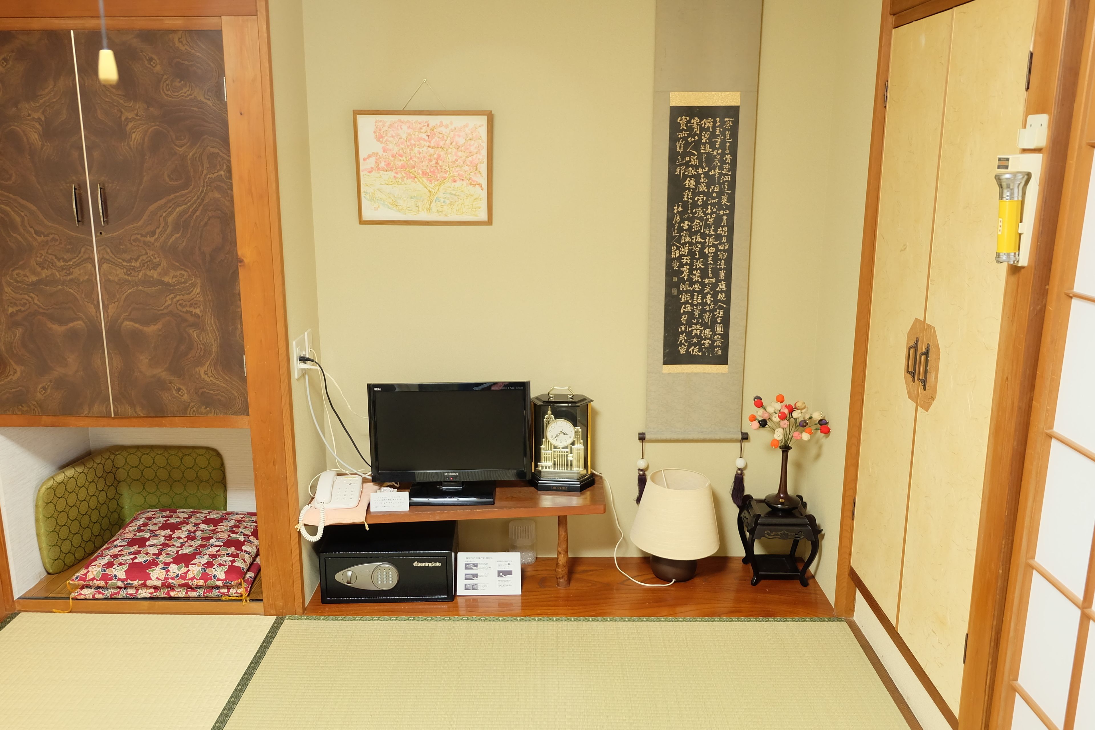 A traditional Japanese room with tatami flooring, a futon bed, a television, a clock, a lamp, and a calligraphy scroll.