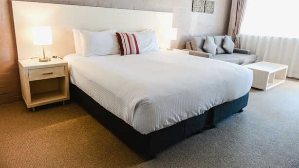 A hotel room with a large bed, a nightstand, a bedside table, a sofa, and a window with curtains. - Balan Village Motel (Bomaderry)