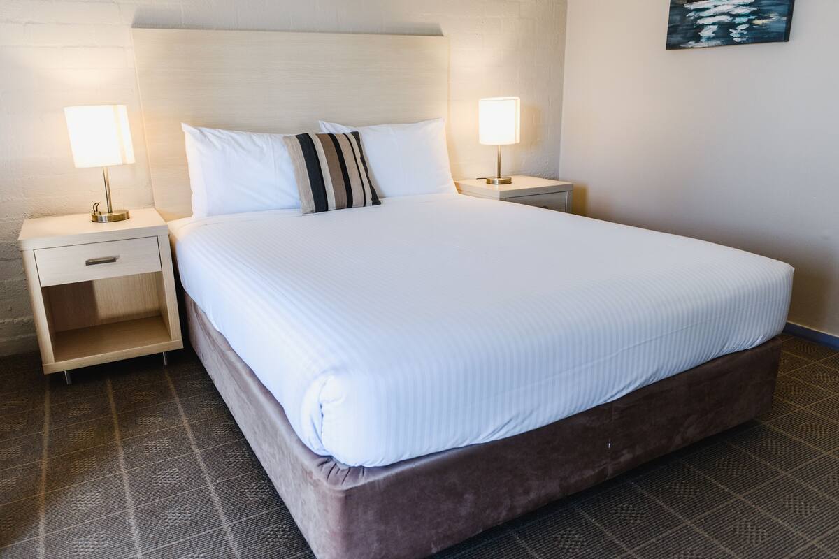 A neatly made bed with white linens and a striped pillow, two bedside lamps, a wooden nightstand with a drawer, and a patterned carpet.
