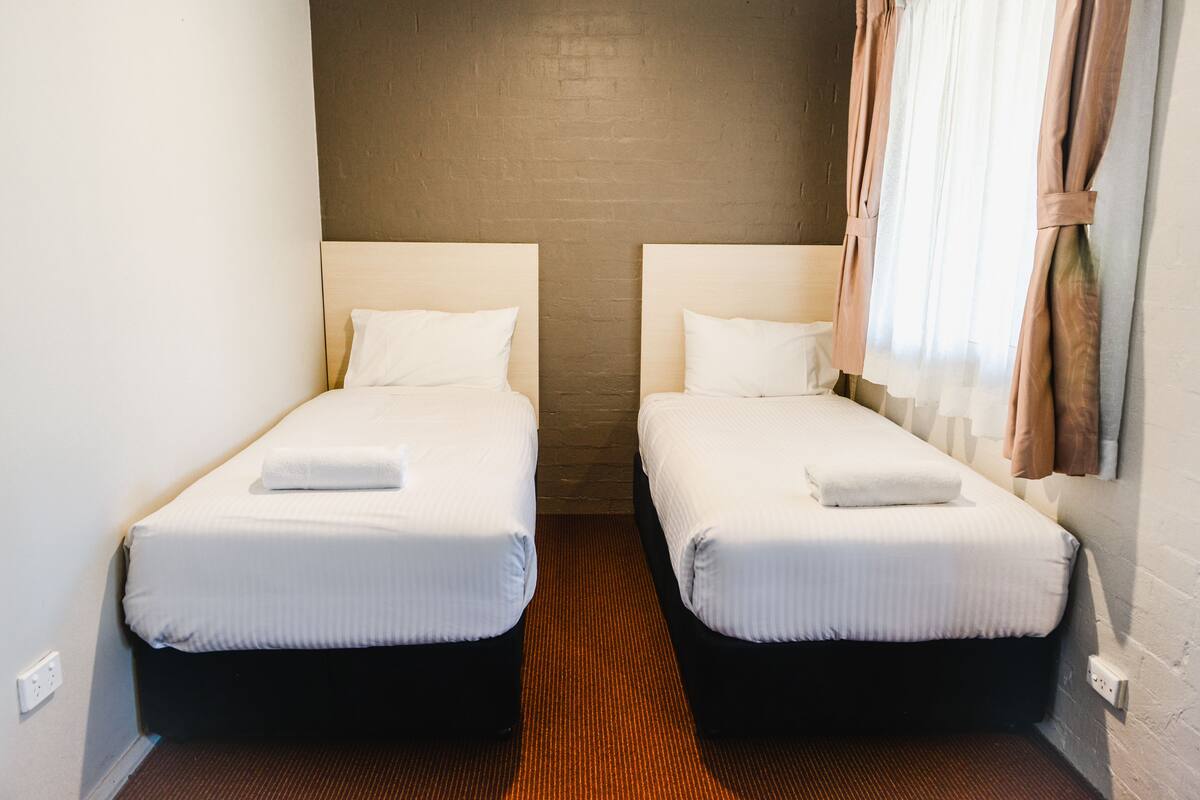A hotel room with two single beds, a brick wall, and a window with curtains.