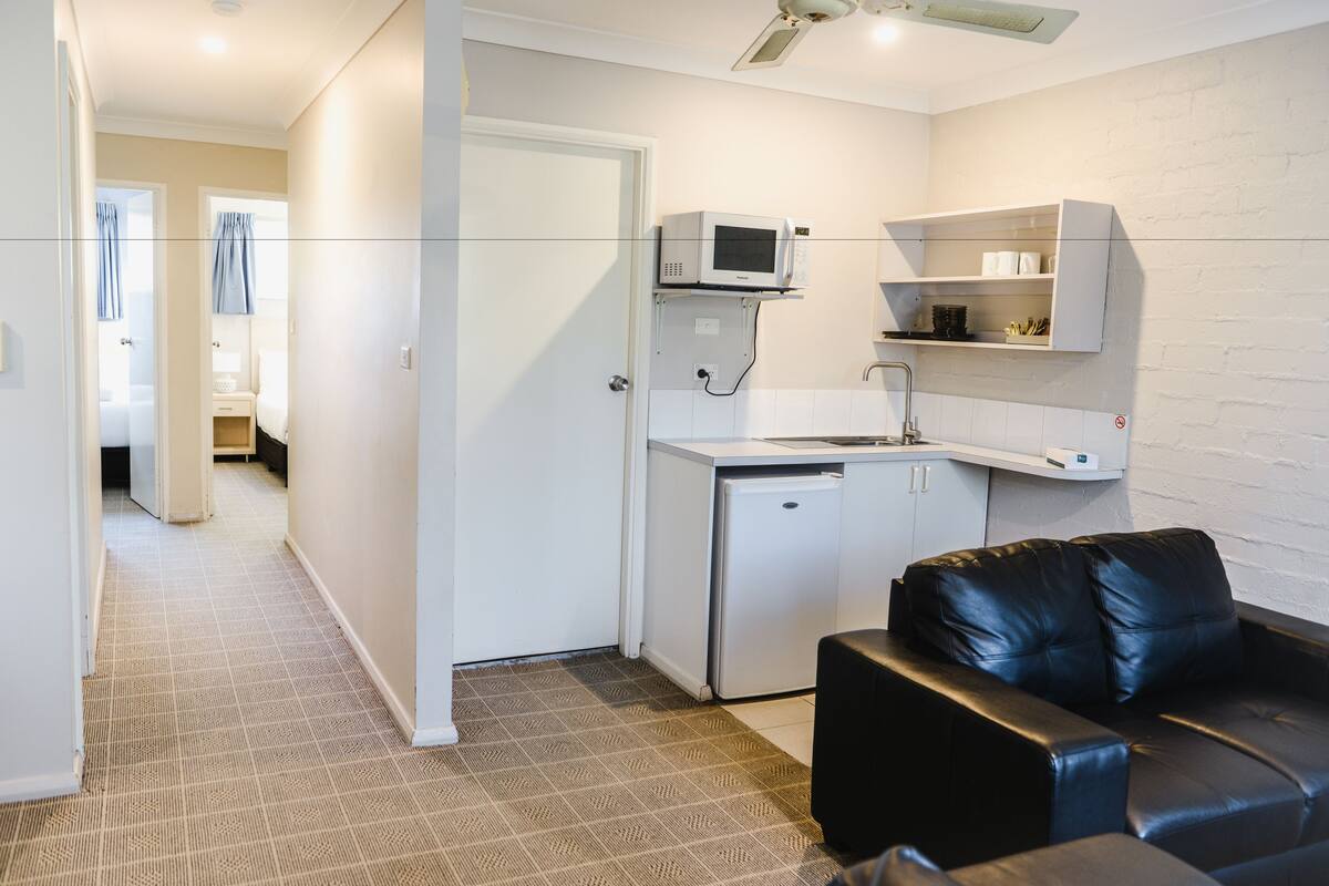 A compact kitchen with a microwave, refrigerator, and white cabinets. There is a black leather sofa in the living area.