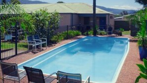 A rectangular swimming pool with lounge chairs and a palm tree, surrounded by a fence and greenery. - Balan Village Motel (Bomaderry)