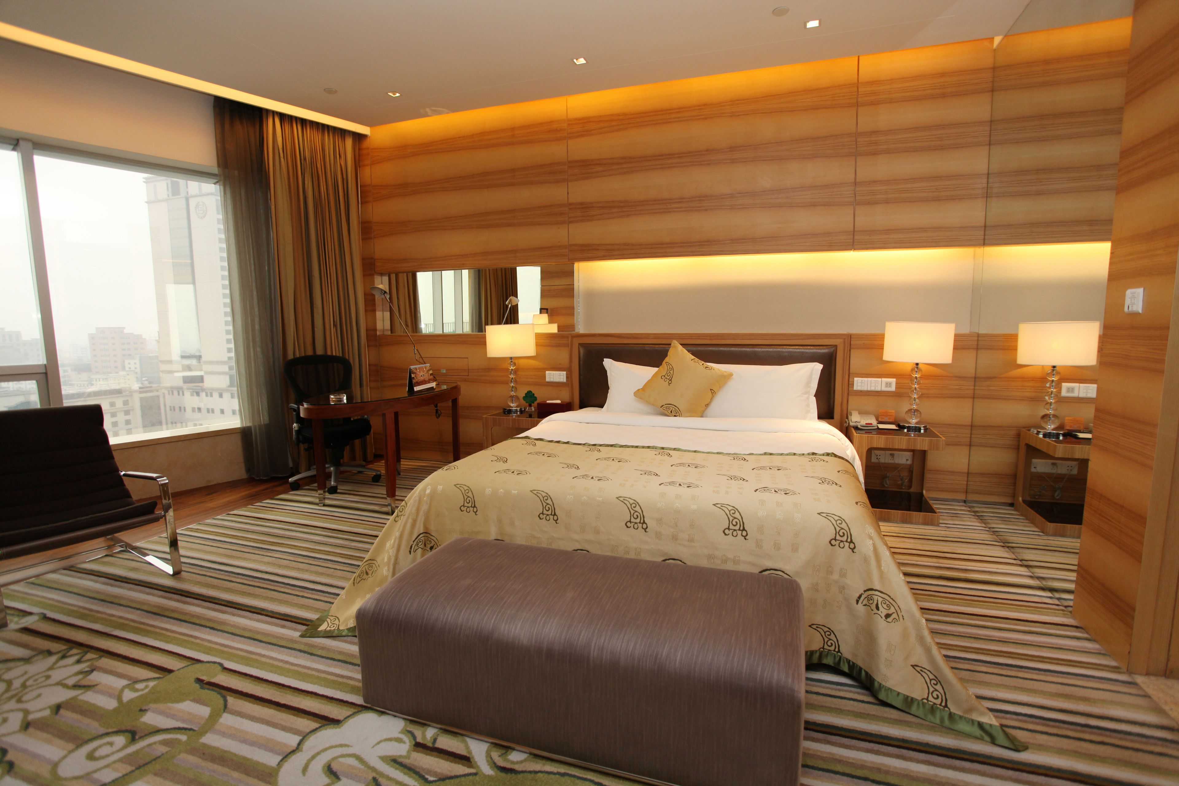 A hotel room with a large bed, two bedside lamps, a desk with a chair, a TV, and a large window with curtains.