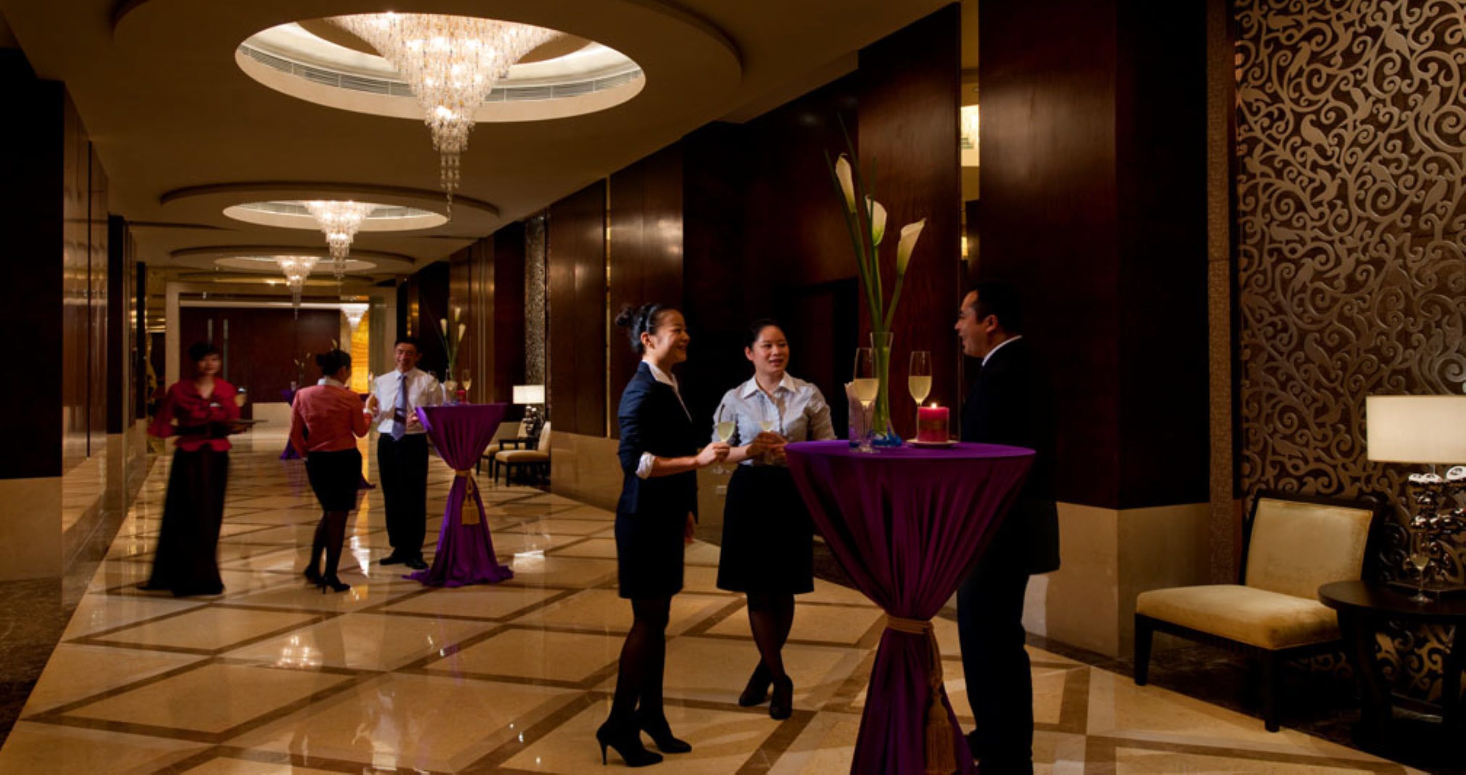 A hotel lobby with a polished marble floor, a grand chandelier, and a reception area with staff.