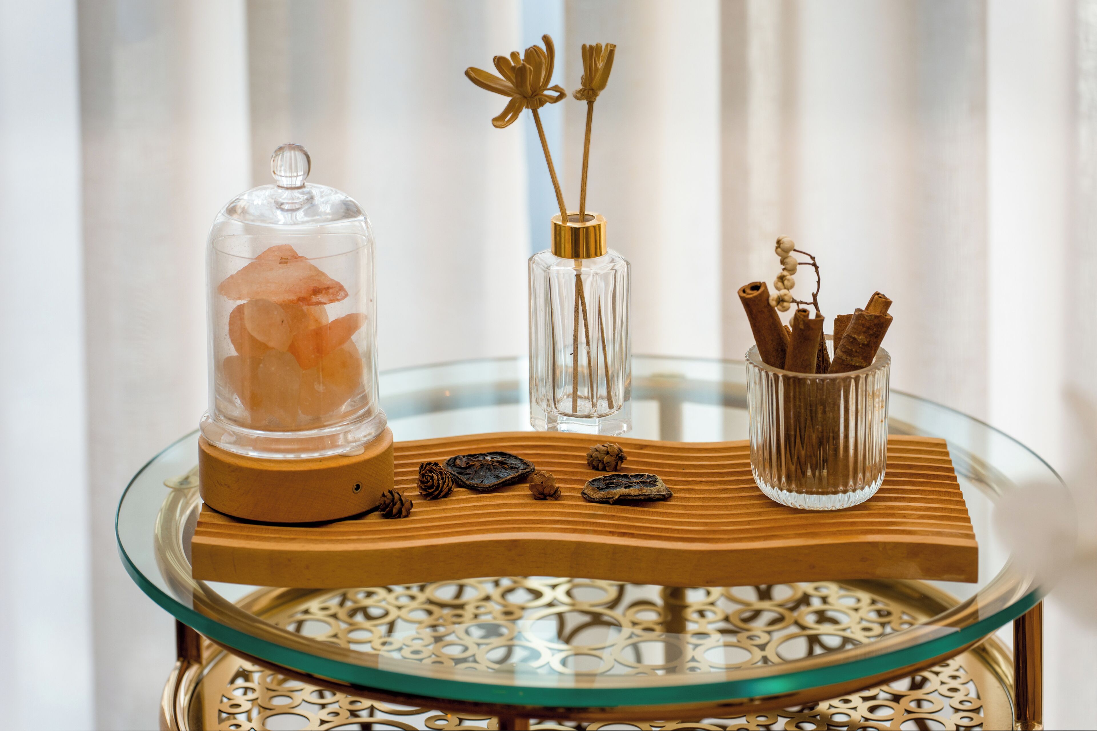 A glass table with a wooden base and decorative items including a glass dome, a clear vase with dried flowers, and a container with cinnamon sticks.