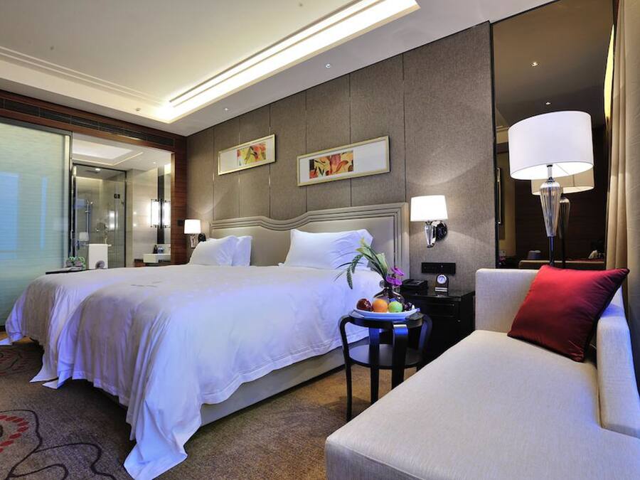 Habitación de hotel con una cama grande, una mesita de noche con lámpara, una silla y una pequeña mesa con fruta.
