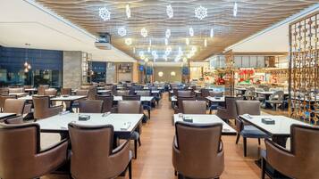 Een ruime restaurantzaal met meerdere eettafels en stoelen, een modern plafond en een decoratieve wand aan de rechterkant.