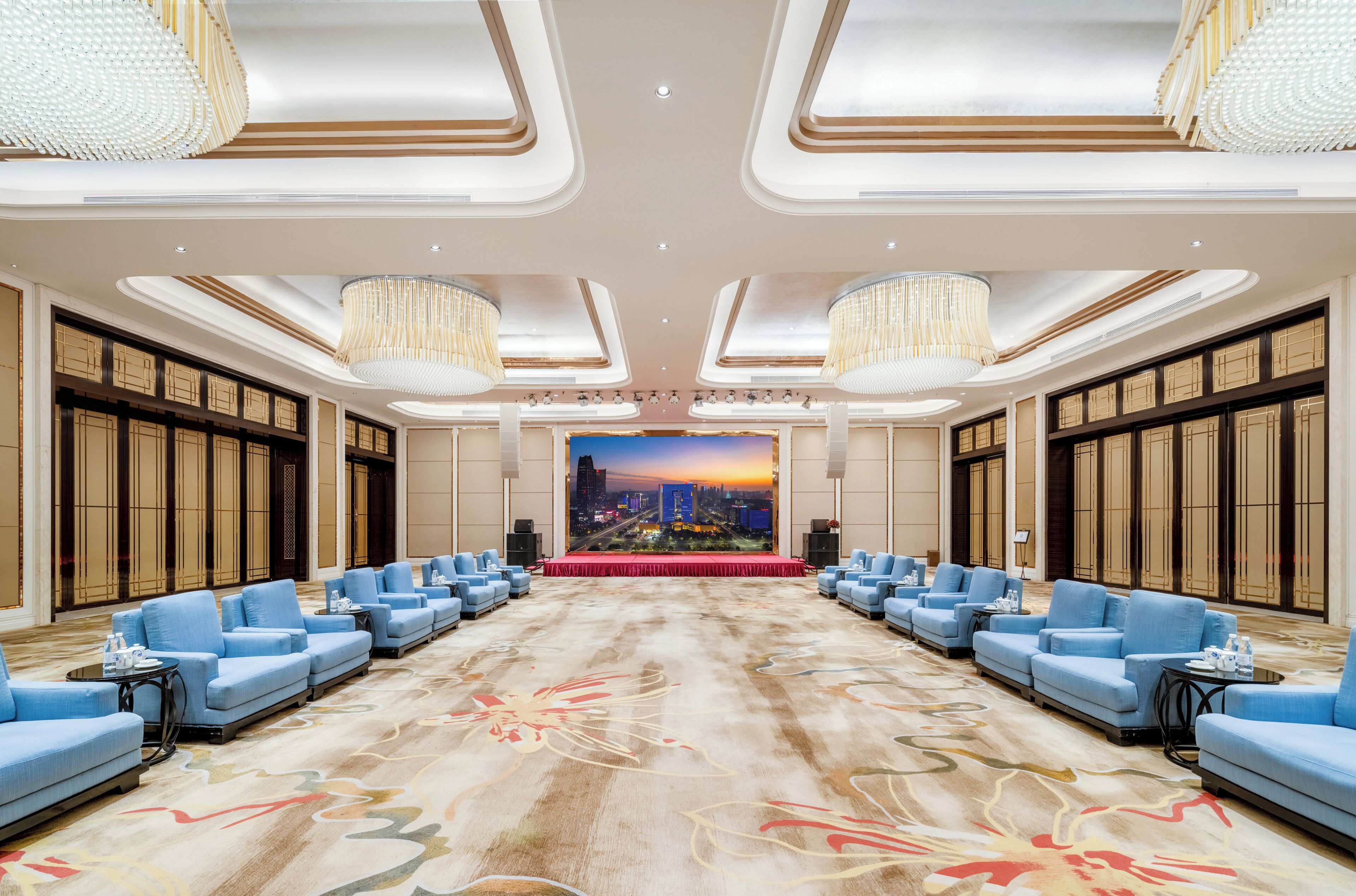 A spacious conference room with a red carpet, blue sofas arranged in rows, and a cityscape backdrop.
