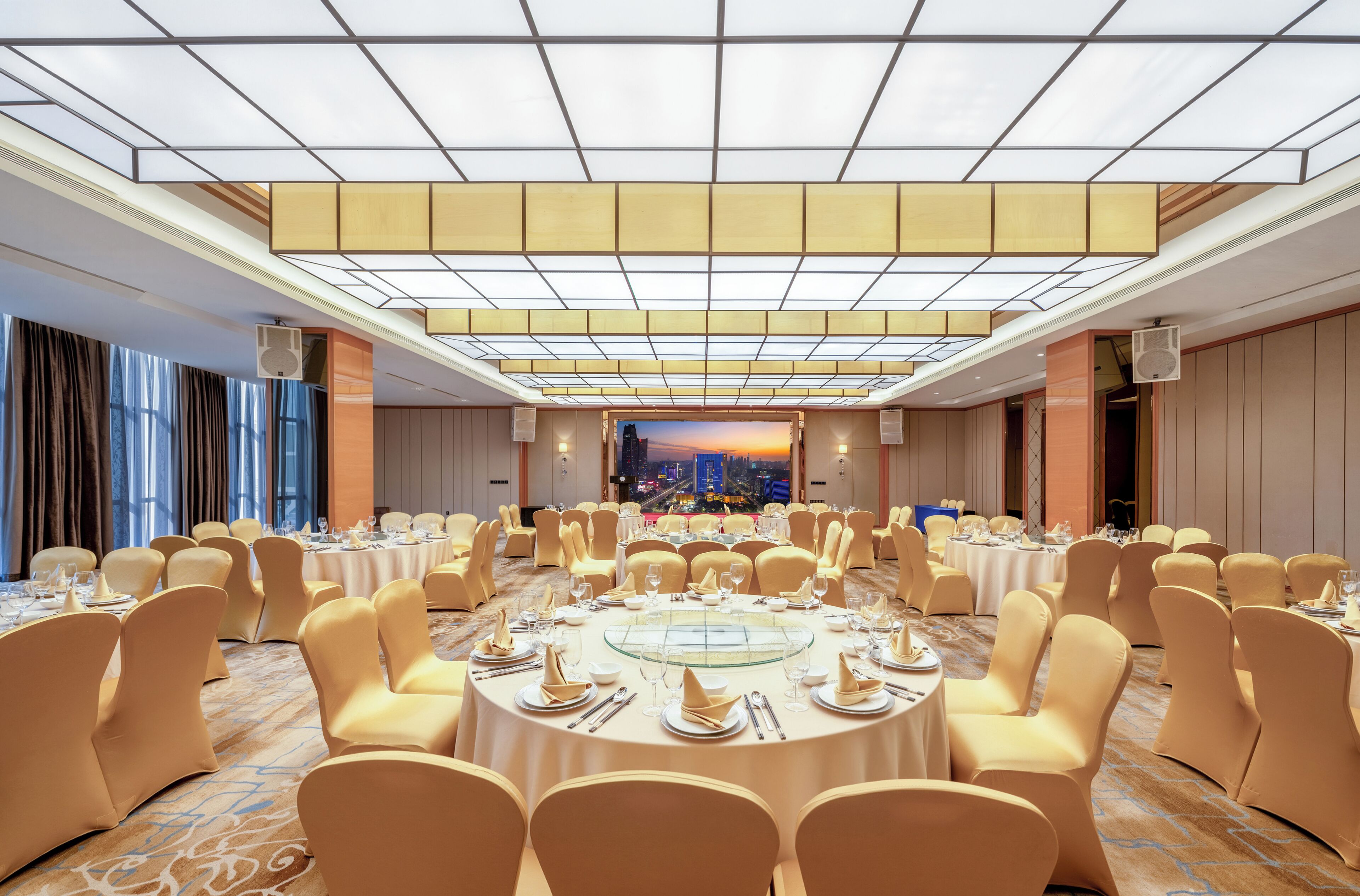 A banquet hall with round tables set for dining, large windows, and a cityscape view.