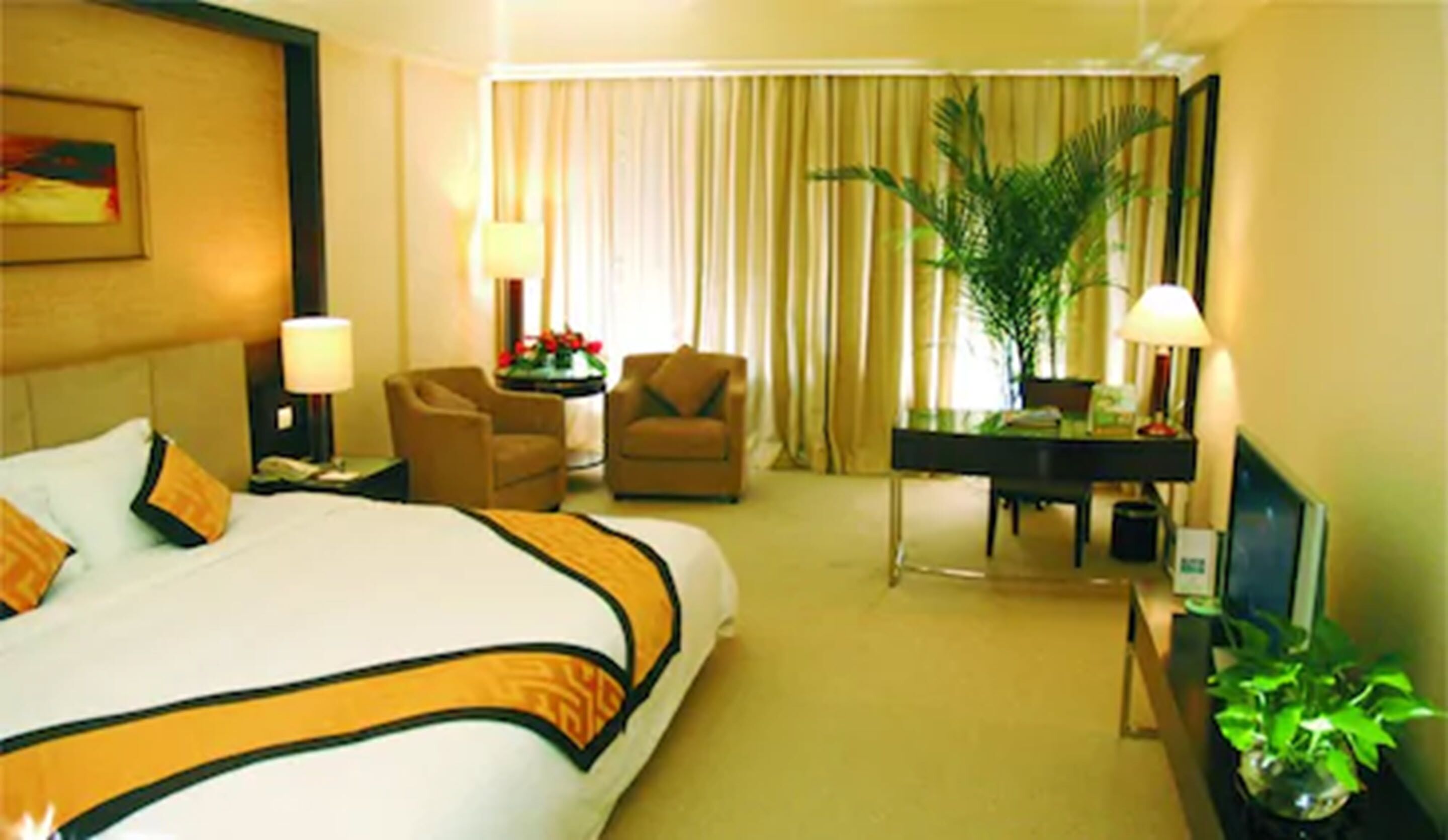 A hotel room with a large bed, two armchairs, a desk, a television, and a potted plant.