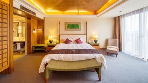 Une chambre d’hôtel avec un plafond en bois, un grand lit avec une banquette, deux tables de chevet avec des lampes, une chaise et une grande fenêtre avec des rideaux.