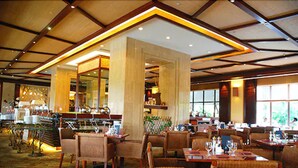 Un restaurant spacieux avec de hauts plafonds, des poutres en bois et une atmosphère chaleureuse. Les tables sont disposées avec des chaises, et le sol est recouvert d’un tapis à motifs.