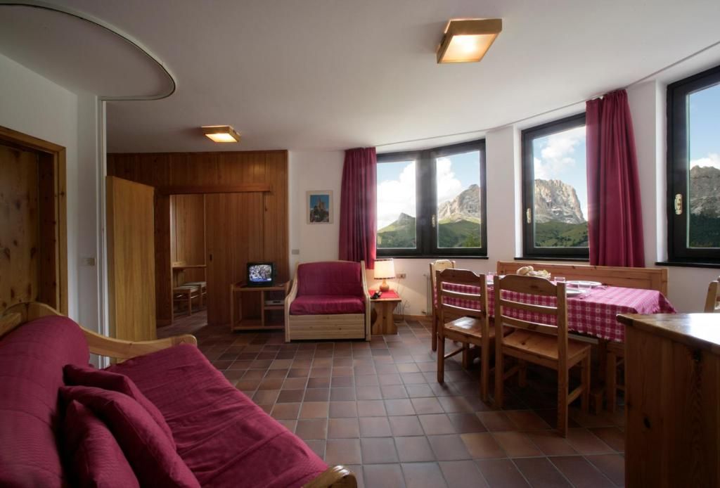 A cozy room with a bed, a desk, a chair, and a table with a checkered tablecloth. There are windows with curtains, a television, and a view of mountains.
