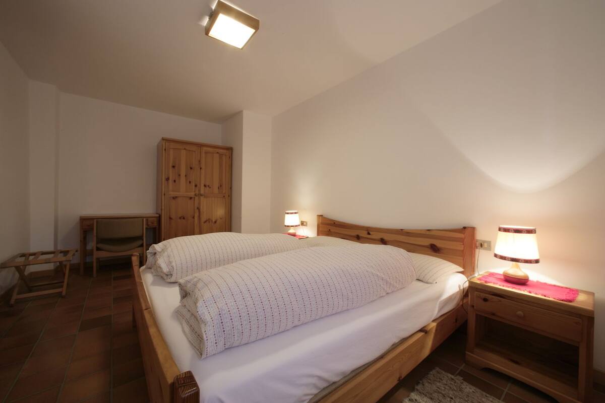 Un dormitorio con una cama de madera, dos mesitas de noche con lámparas, un armario de madera y una pequeña silla de madera.