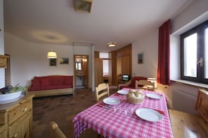 A dining area with a table set for two, a wooden sofa, and a kitchen area visible in the background.