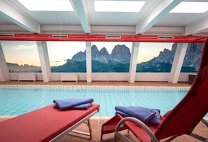 A swimming pool with red lounge chairs and towels, with a mountain view through the windows. - Hotel Garni Gonzaga (Canazei)