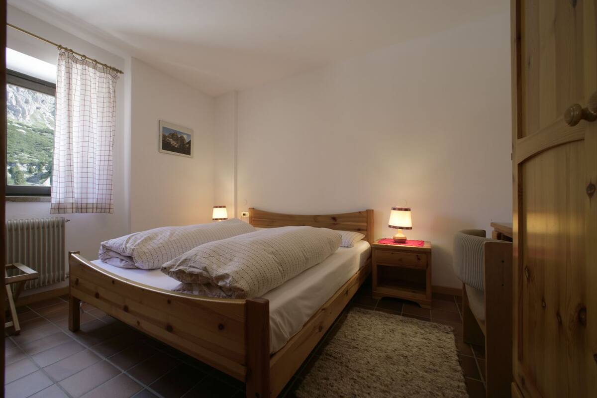 Un dormitorio con cama de madera, dos lámparas de noche, un radiador y una ventana con cortinas.