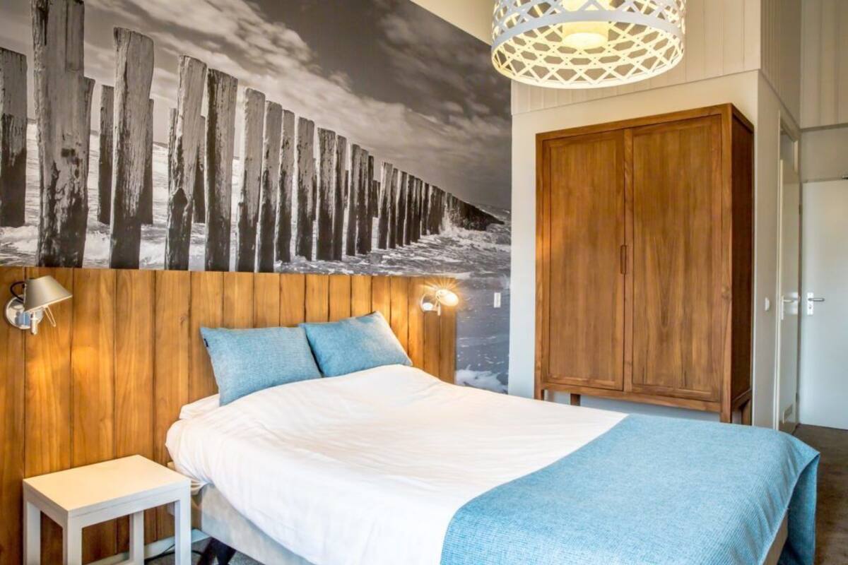 Un dormitorio con cabecera de madera, cama con ropa de cama blanca y almohadas azules, mesita de noche, armario de madera y un mural en la pared con una escena de bosque.