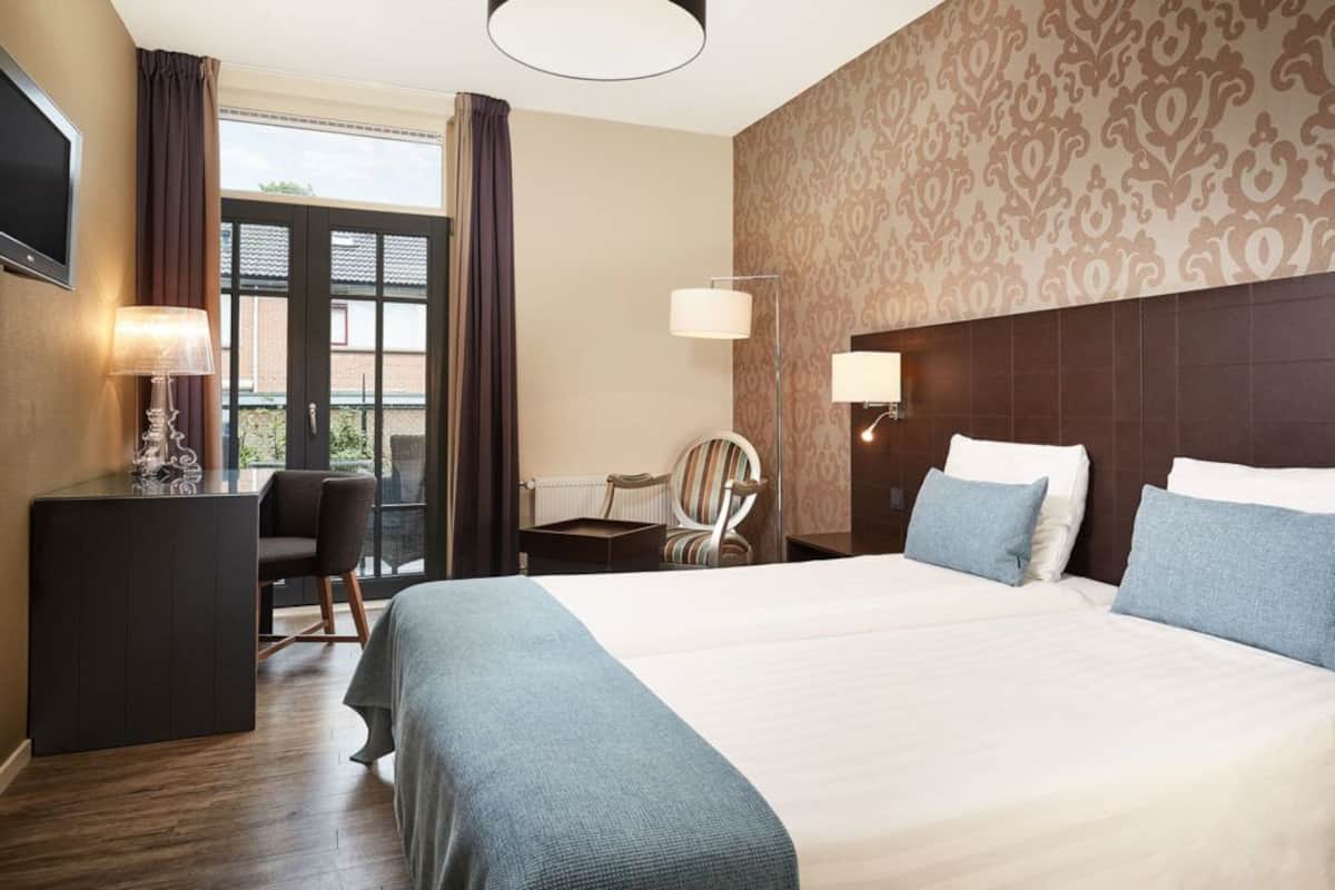 Habitación de hotel con una cama grande, un escritorio con lámpara, una silla y vistas al exterior a través de una ventana.