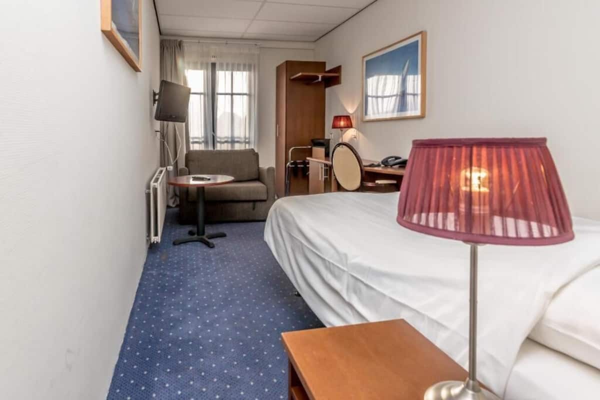 Habitación de hotel con cama, escritorio, silla, lámpara y ventana con cortinas.