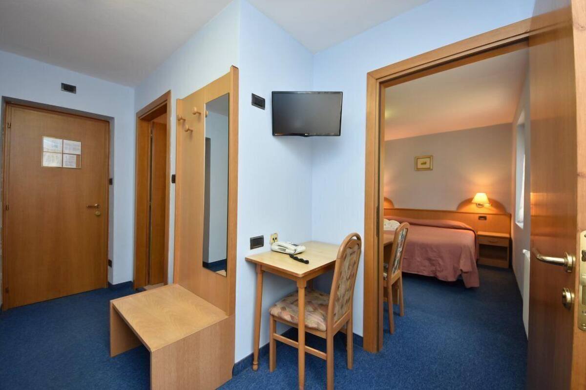 A compact hotel room with a wooden desk, a chair, a bed with a pink bedspread, a television mounted on the wall, and a door leading to another room.