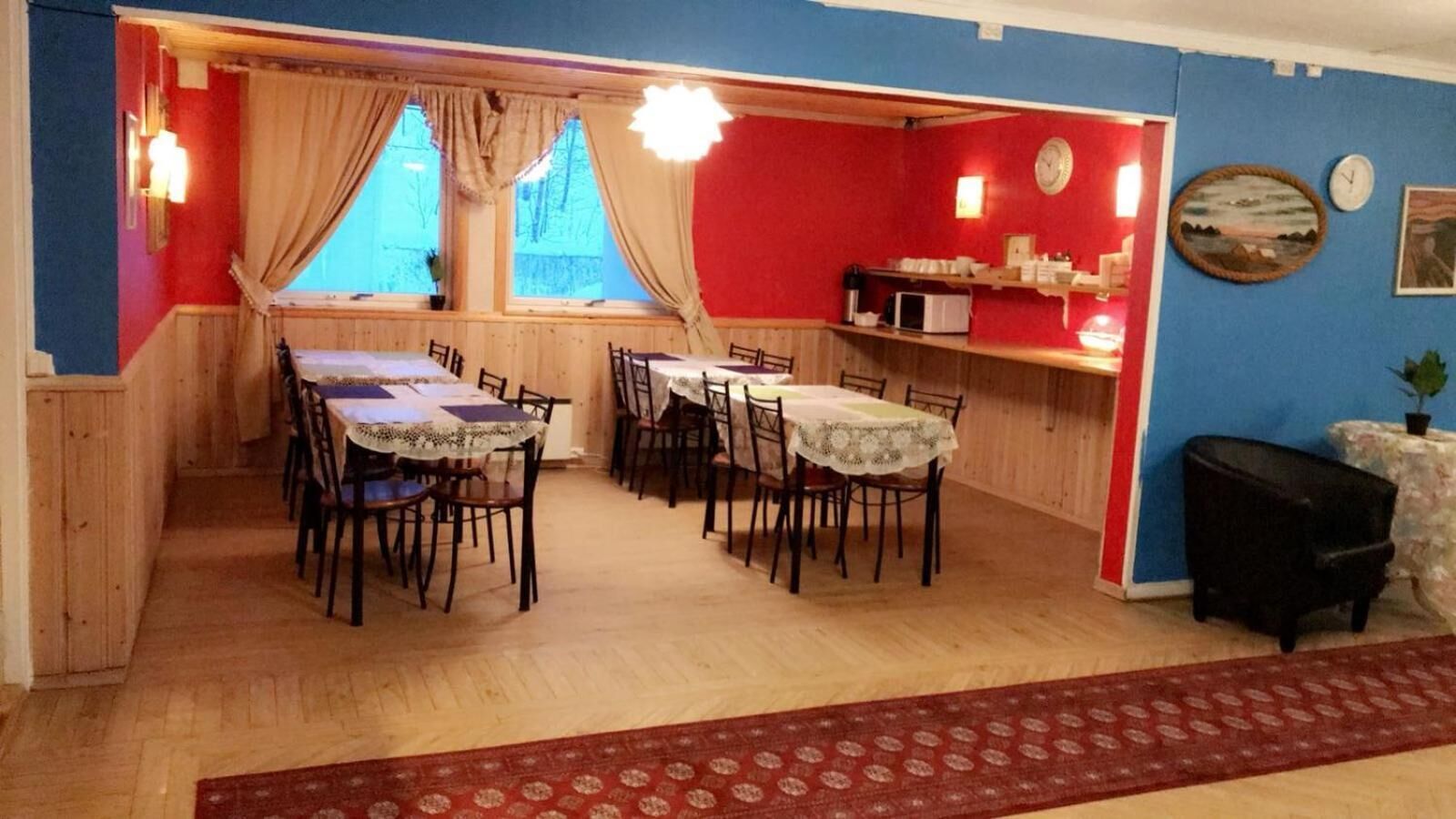 A dining area with wooden tables and chairs, a kitchen area with a microwave, and a red wall with a clock and artwork.