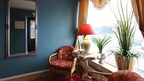 A room with a blue wall, a wooden chair with floral upholstery, a red lamp, and a large potted plant. - Barents Frokosthotell (South Varanger)