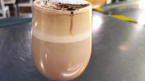 Una taza de bebida de chocolate con espuma, sobre una mesa en un café con vistas al exterior, donde se ven plantas.