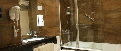 Un baño con ducha de vidrio, inodoro blanco, lavamanos con espejo y un secador de pelo en la pared.