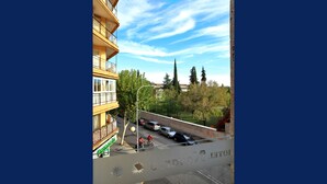 Un edificio de varios pisos con balcones, una calle con coches y un ciclista, y árboles al fondo.