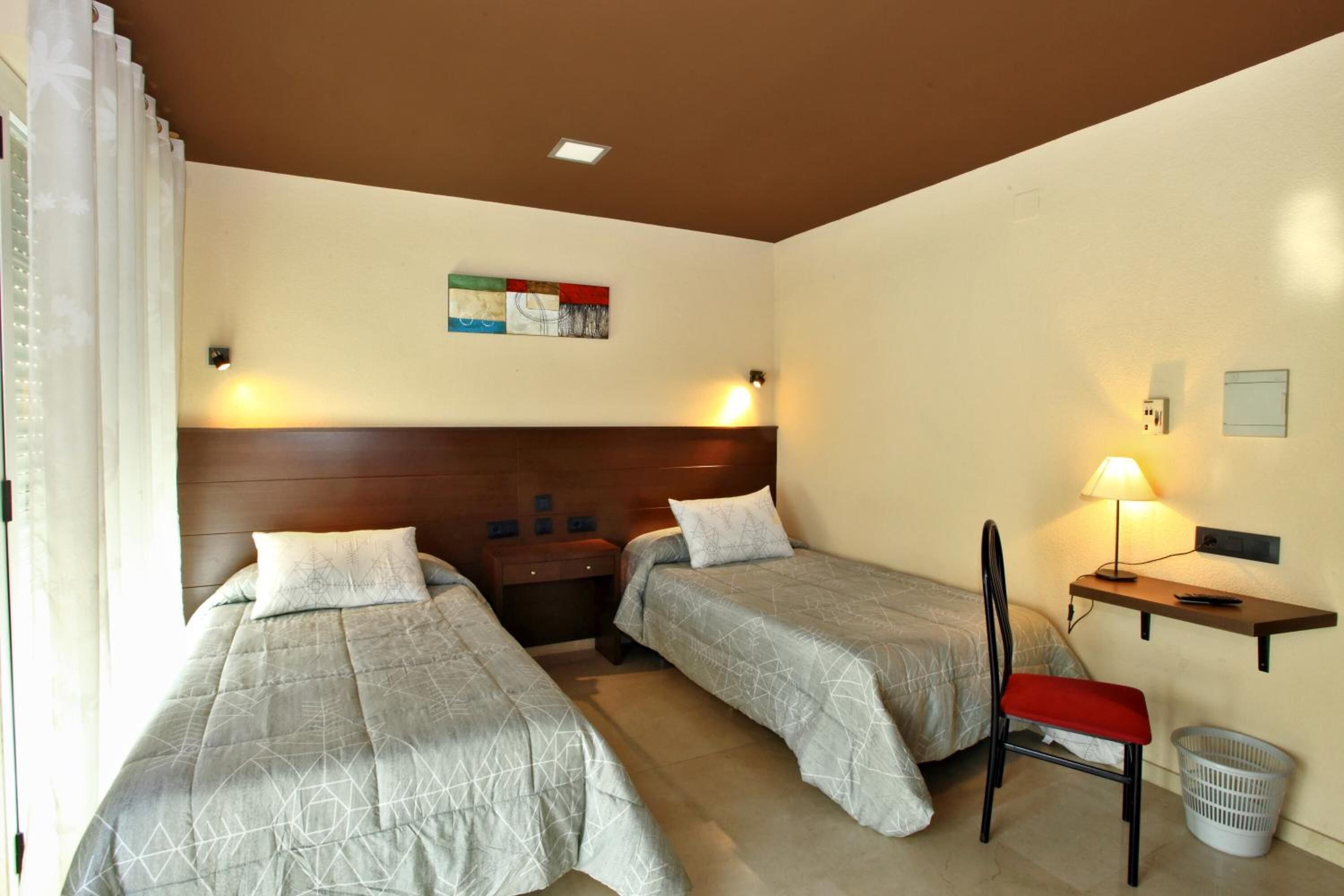 Ein Hotelzimmer mit zwei Einzelbetten, einem Nachttisch, einem Sessel, einem Schreibtisch und einer Wandlampe.